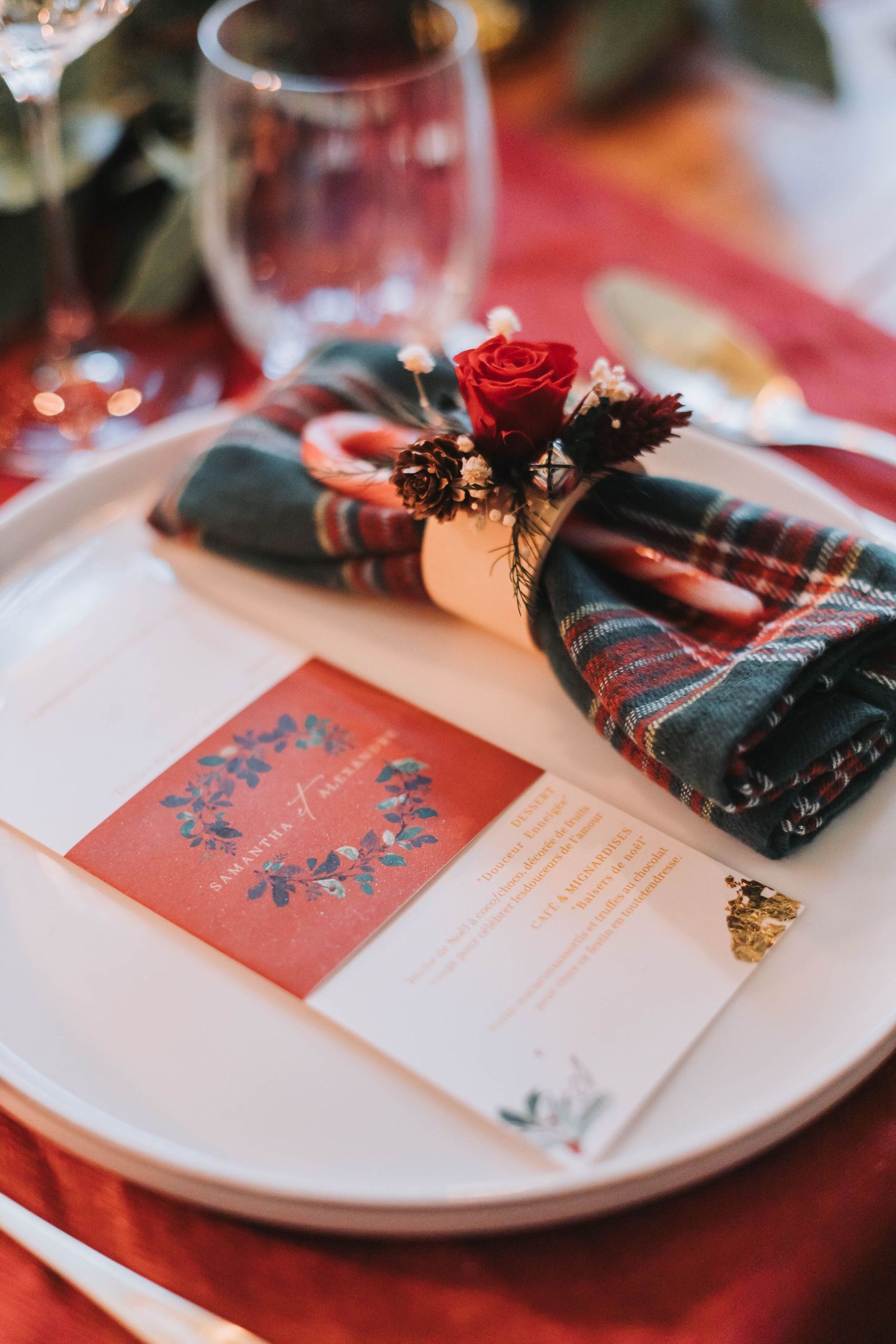Elegant table setting with plaid napkin, red rose and wedding menu card on white plate with burgundy tablecloth.