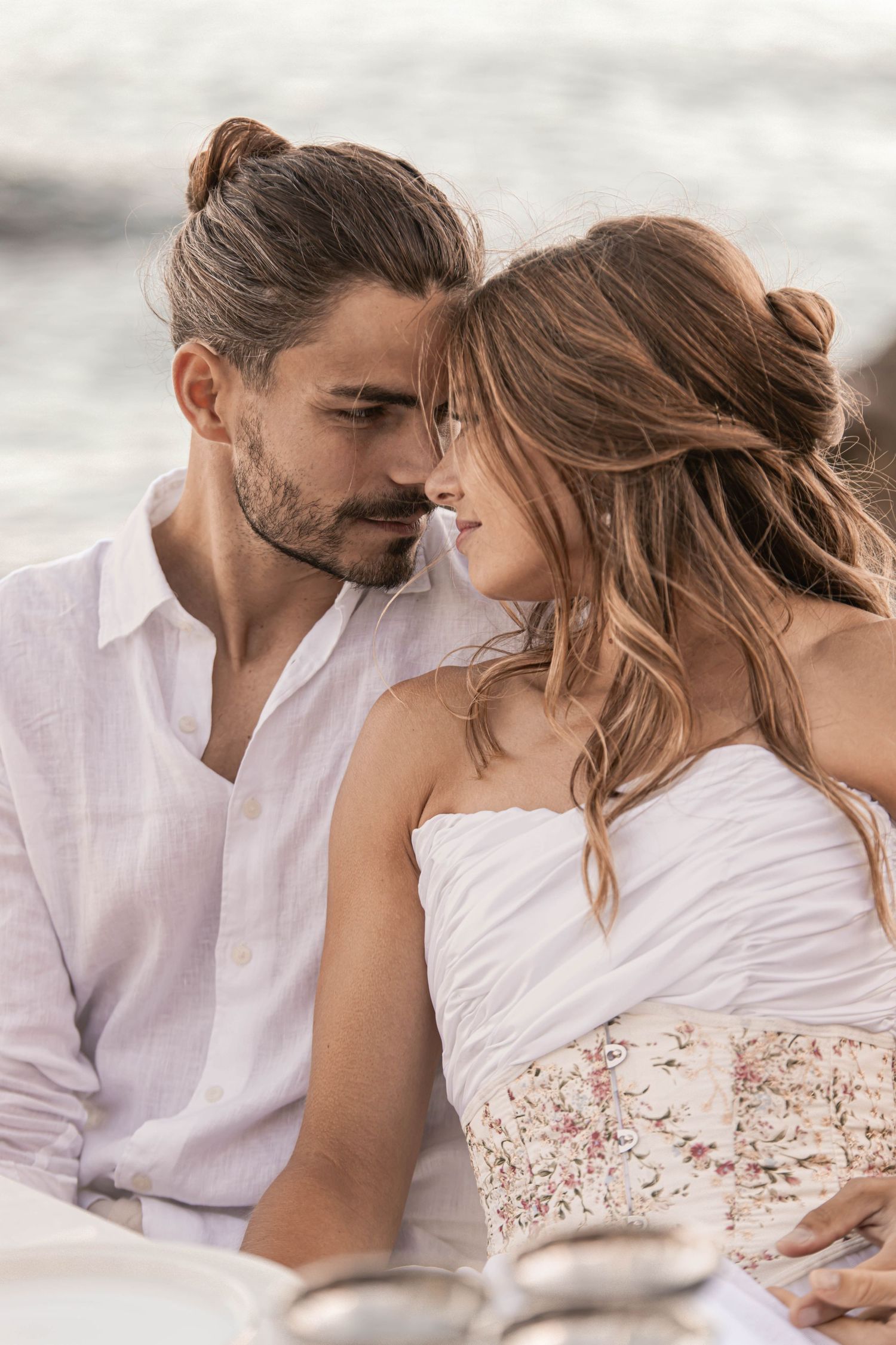 Intimate couple in white outfits sharing a tender moment near the ocean at a beach wedding.