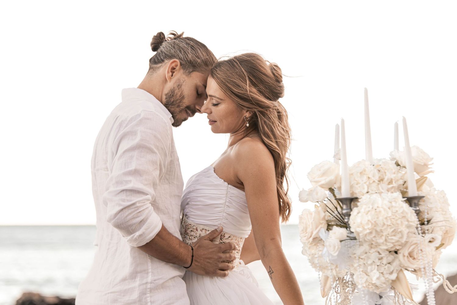 Bride and groom embracing tenderly near white floral arrangements at a seaside wedding.