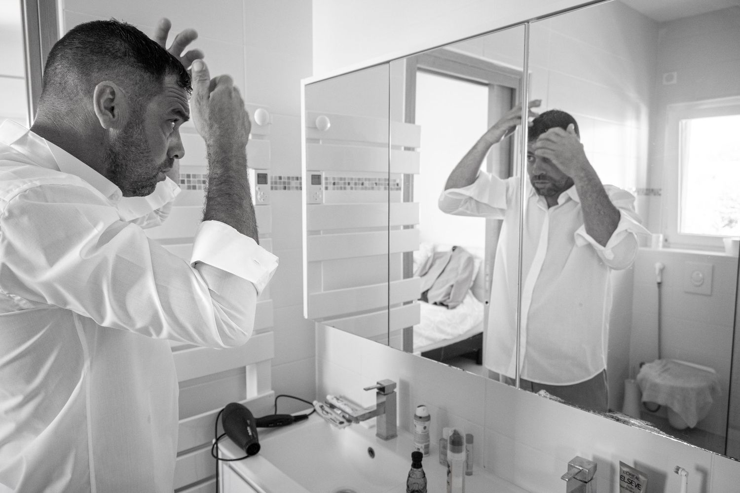 Man in white dress shirt getting ready in front of bathroom mirror on wedding morning.