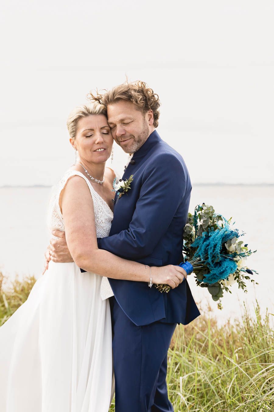Bride holds turquoise bouquet while sharing intimate moment with groom by the seaside.