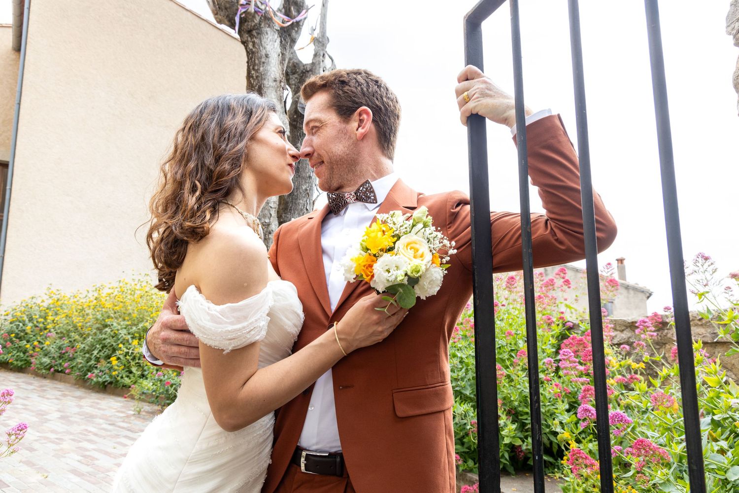 Newlyweds share intimate moment by wrought iron gate with bright floral bouquet.