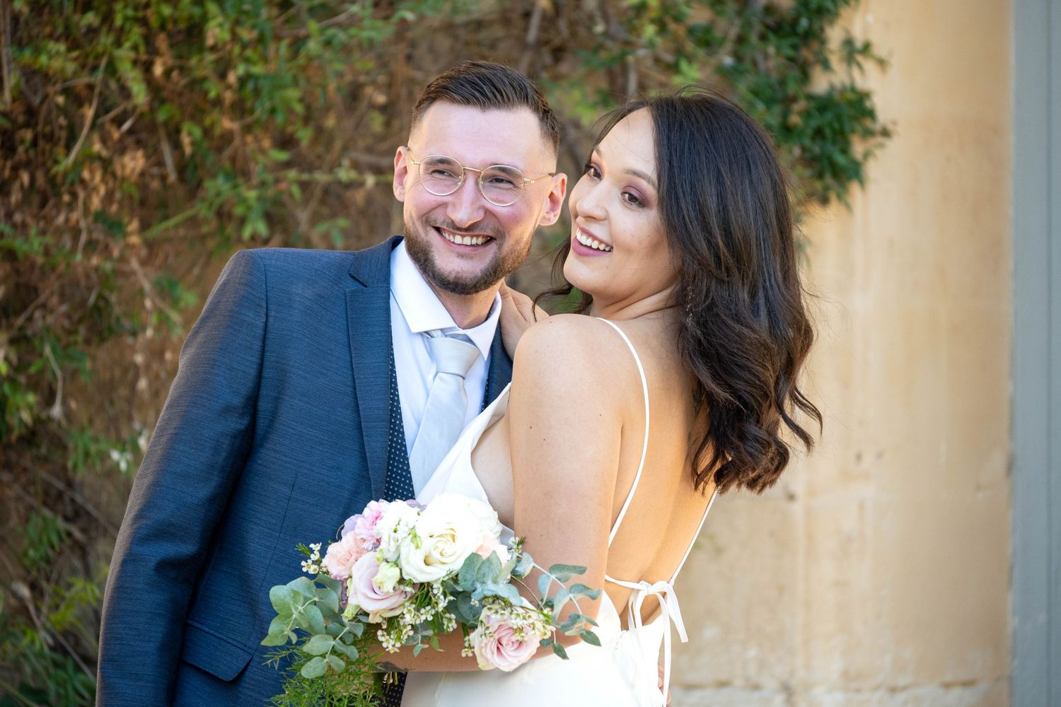 Wedding portrait with couple smiling in front of yellow building and greenery.