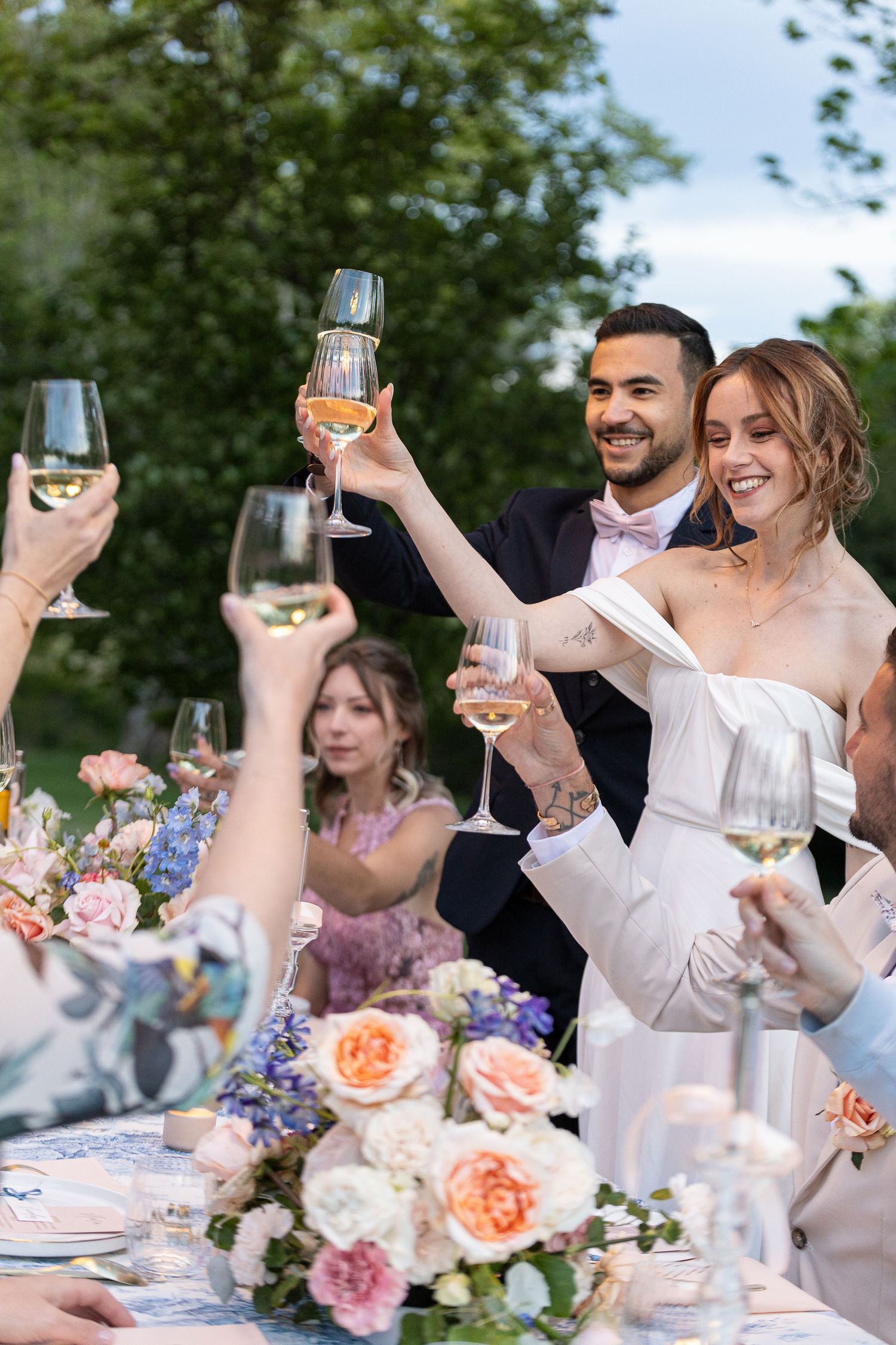 Wedding reception table decorated with pink and purple flowers as guests raise glasses in celebration.