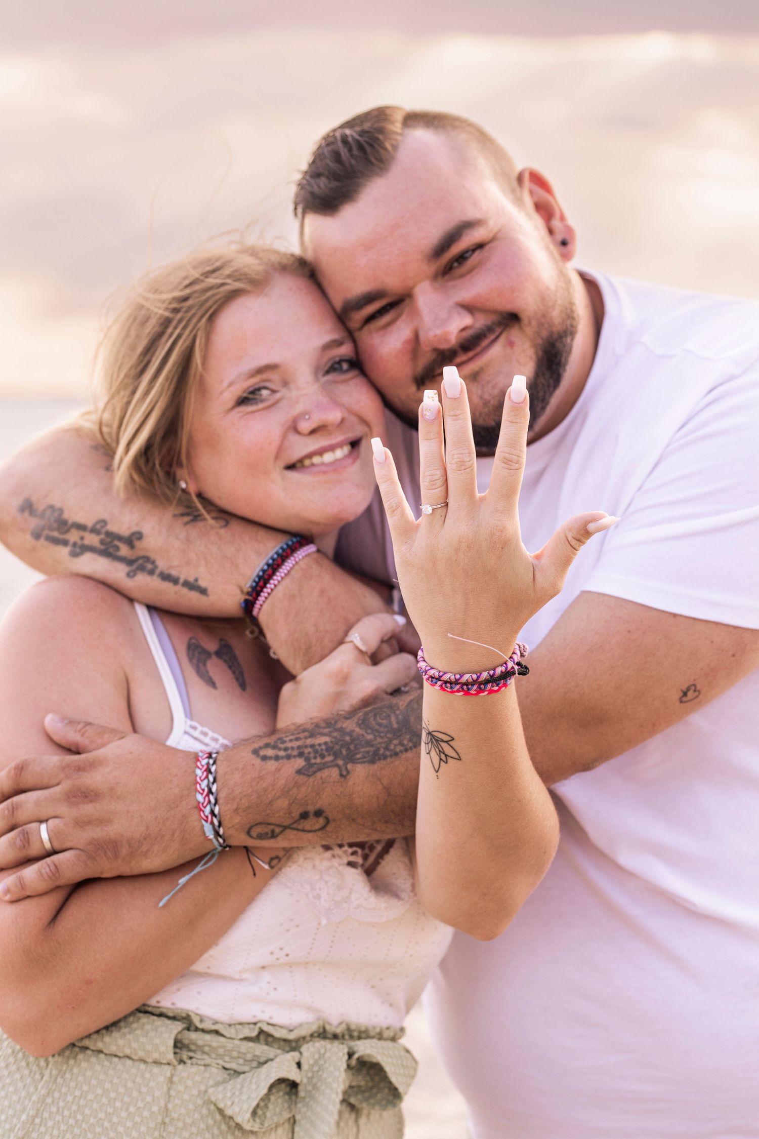 A couple embracing on the beach shows off an engagement ring during their sunset photoshoot.