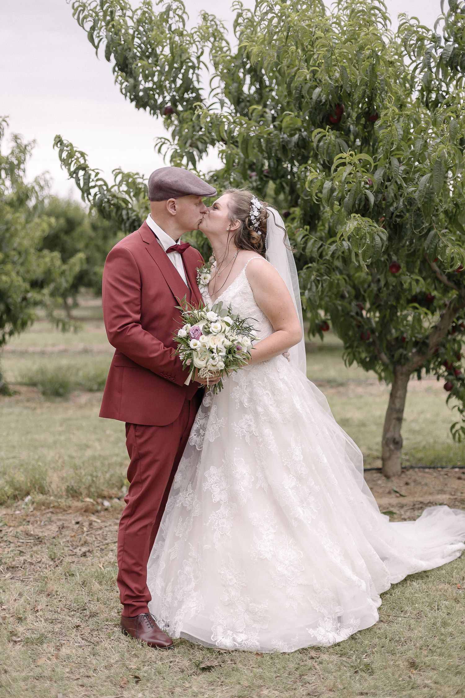 Wedding couple embraces in an orchard wearing elegant white dress and burgundy suit.