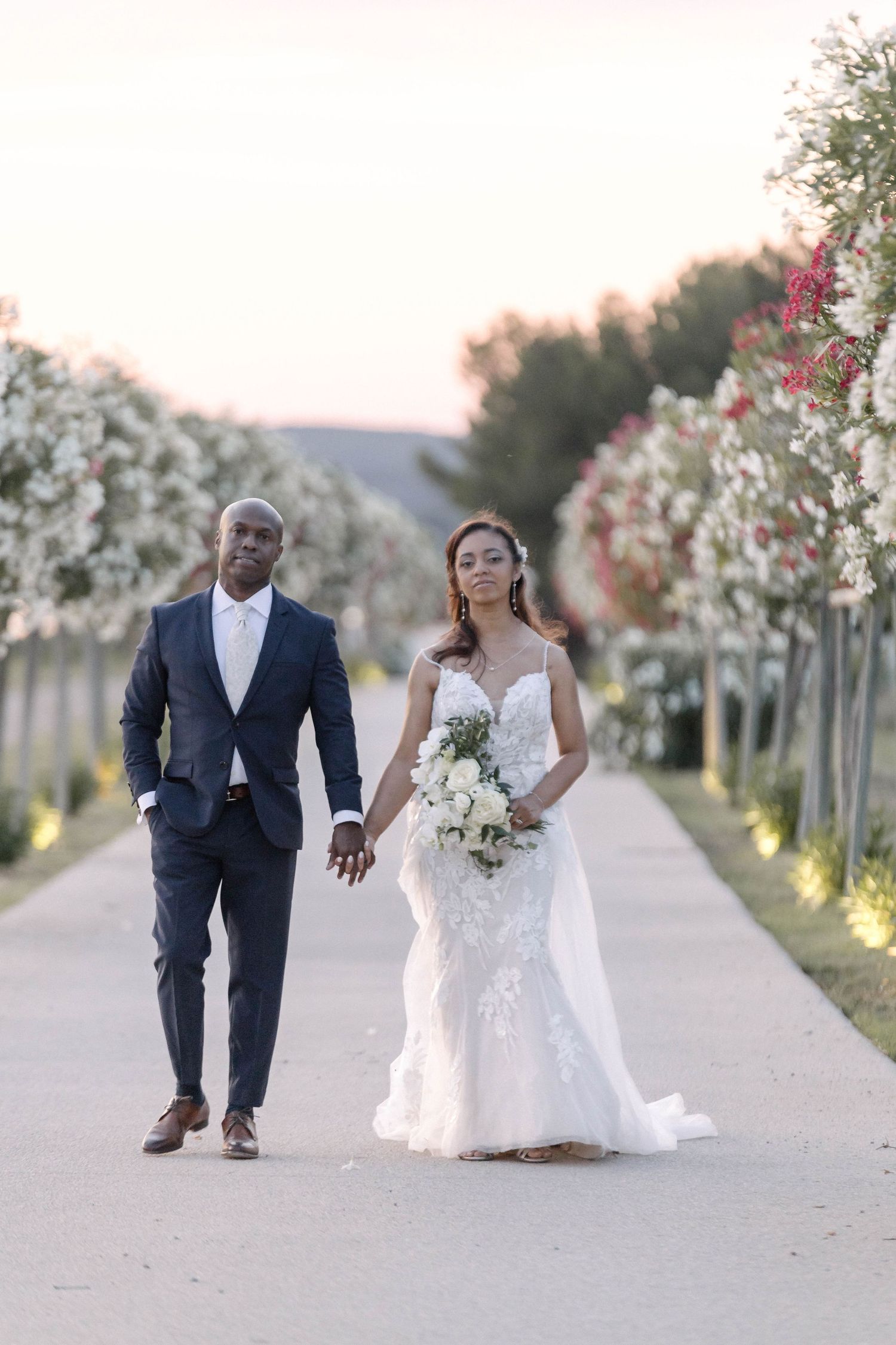 Wedding couple strolls down a flower-lined pathway during their vineyard photoshoot.