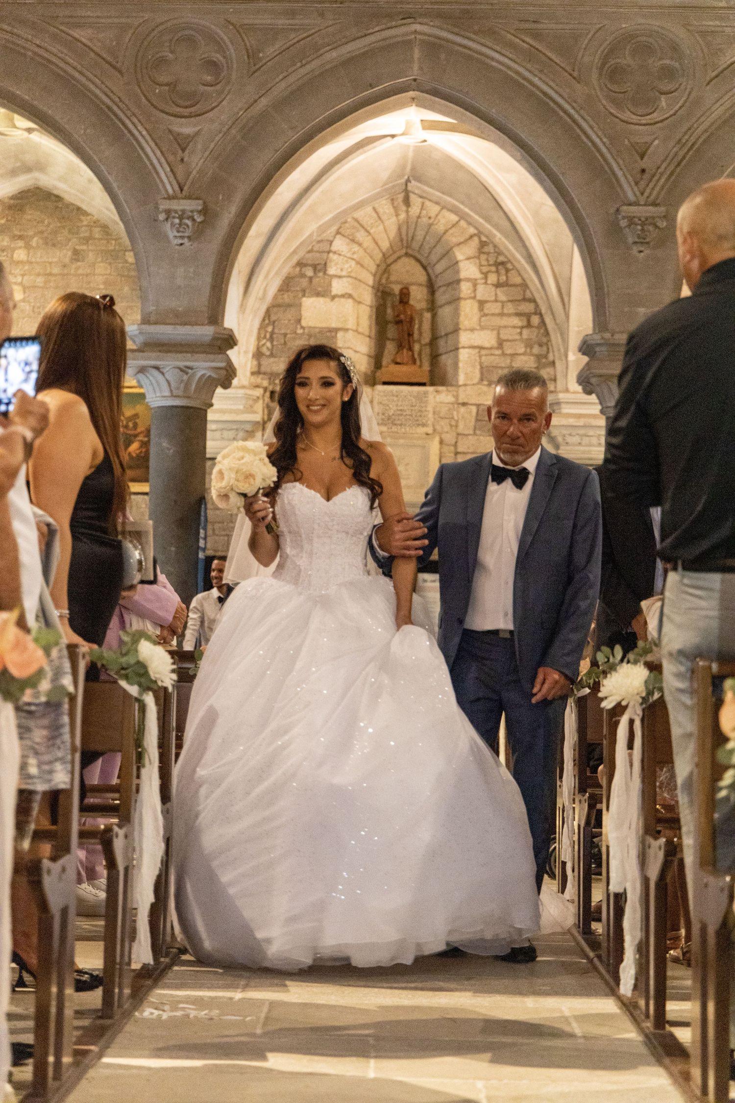 A wedding procession moves through an elegant stone archway with ornate architectural details.