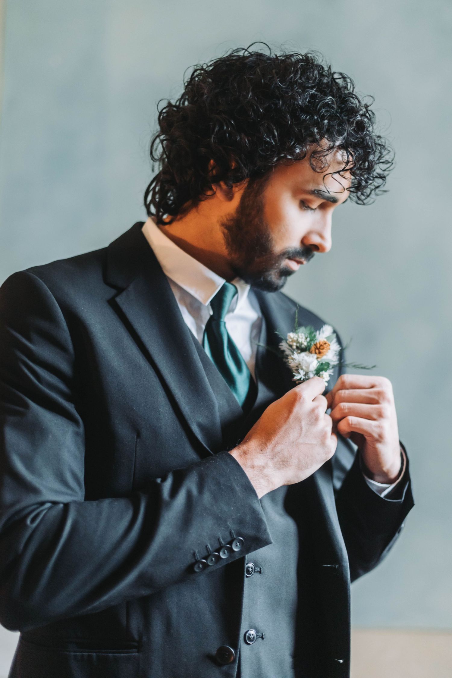 Groom in dark suit with curly hair adjusts boutonniere against light background.