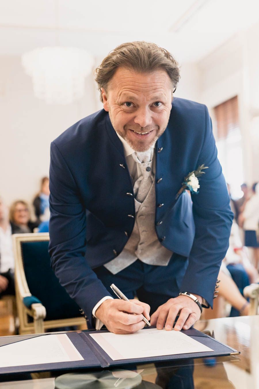Groom in navy suit smiling while signing marriage documents.