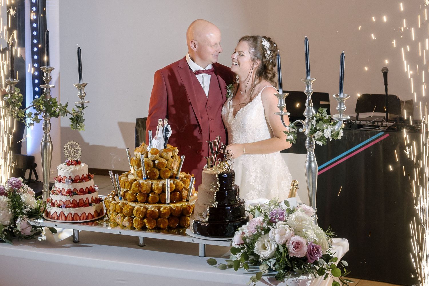 A wedding couple poses by their dessert table featuring a multi-tiered cake with sparklers.