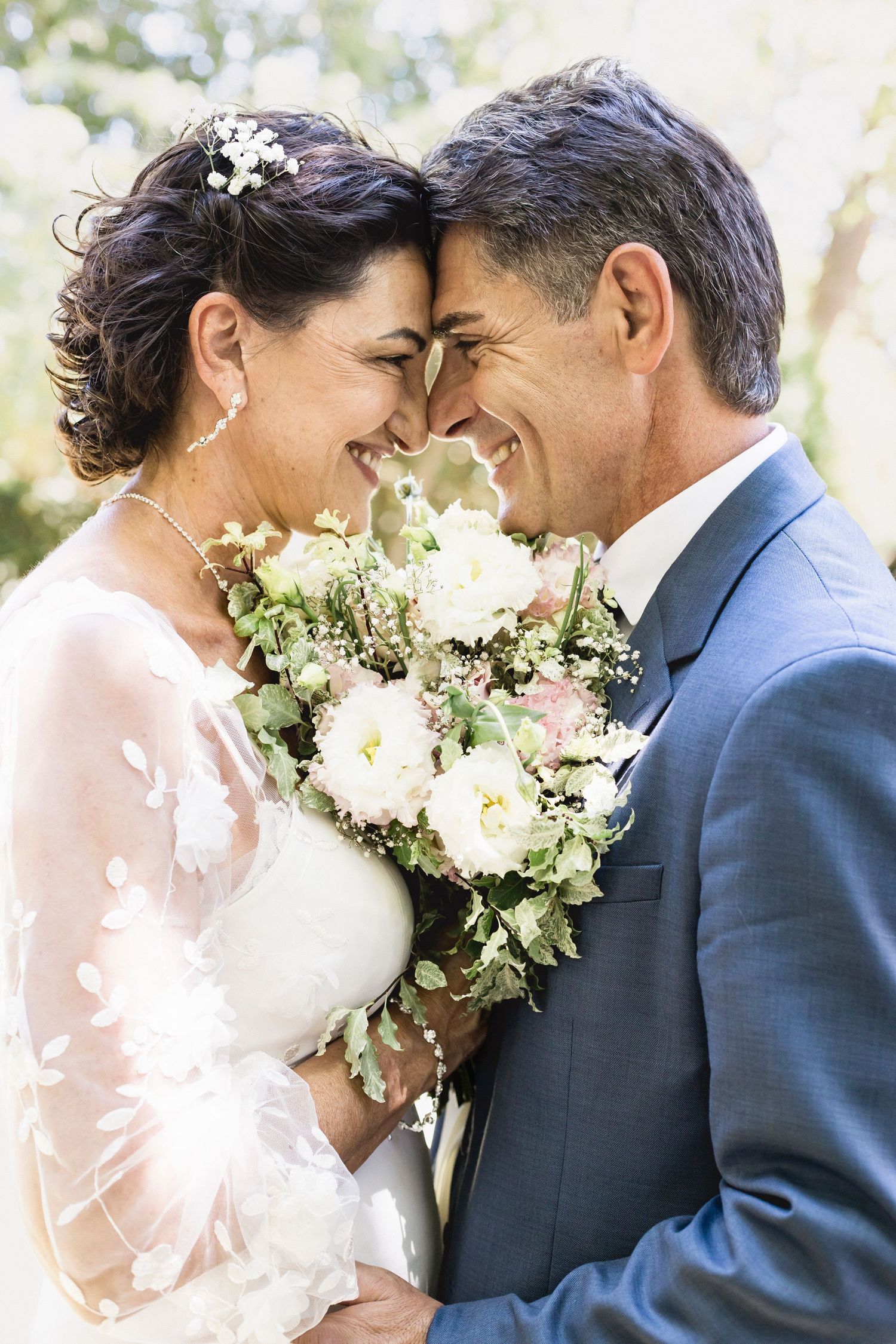 Bride in lace dress and groom in navy suit share romantic moment with elegant white bouquet.