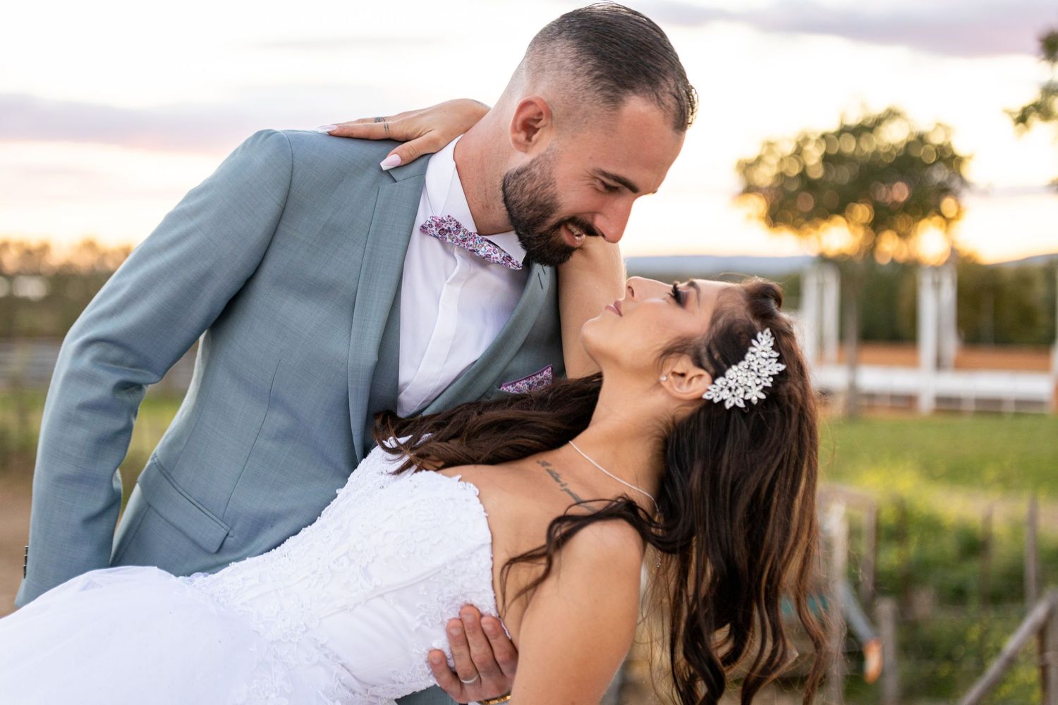 A couple in wedding attire embraces during sunset in a rustic outdoor setting with golden light streaming through trees.