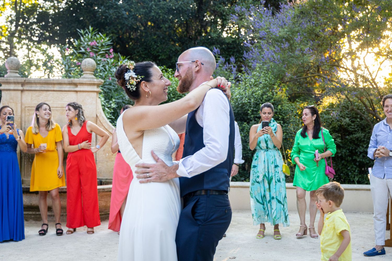 A couple shares their first dance surrounded by guests in colorful dresses in a garden setting.
