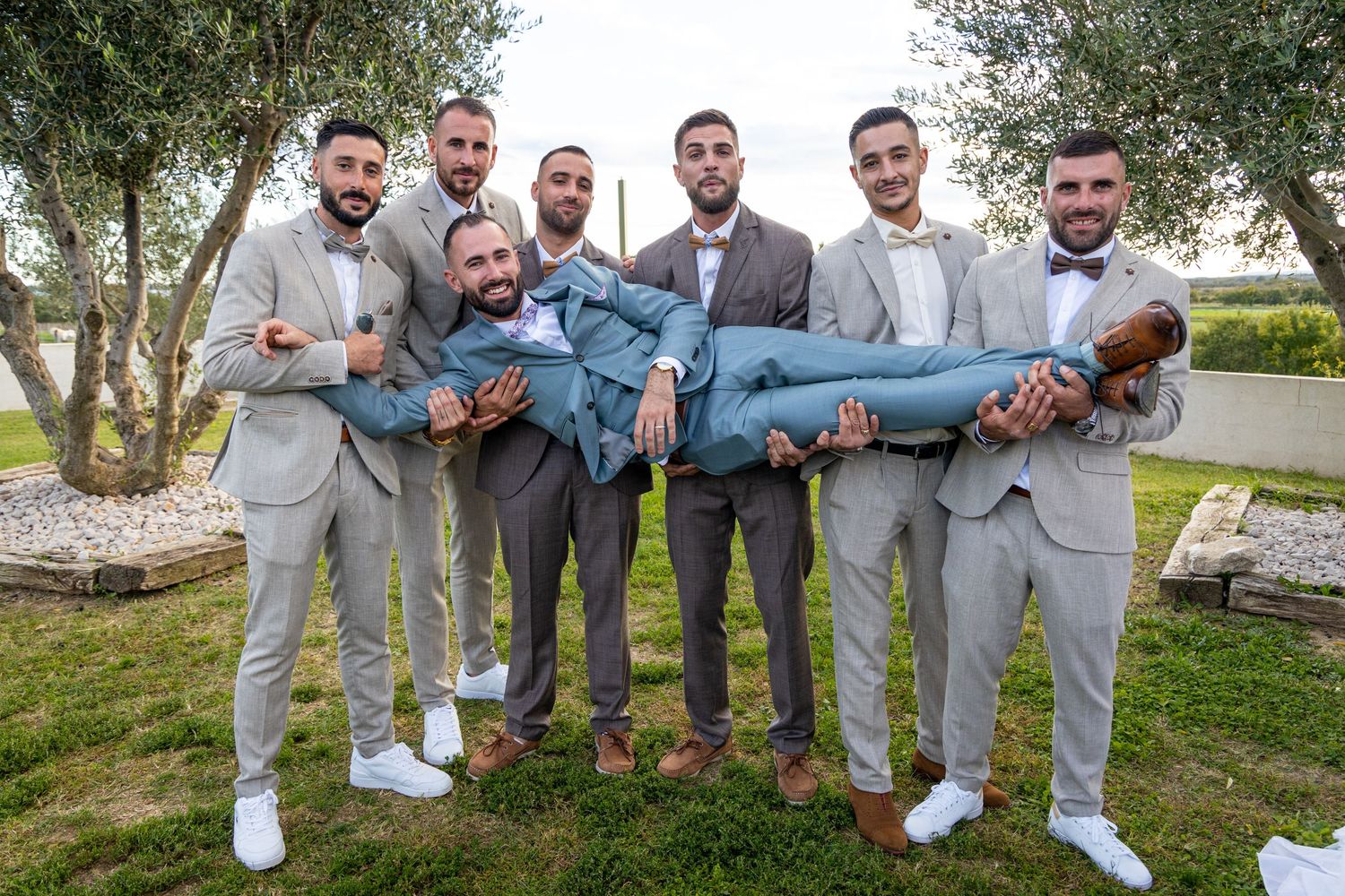 Groomsmen in light grey suits playfully carrying one of their group members.