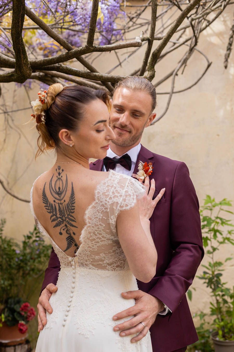 Romantic wedding couple embraces under blooming wisteria in garden setting with white dress and burgundy suit.