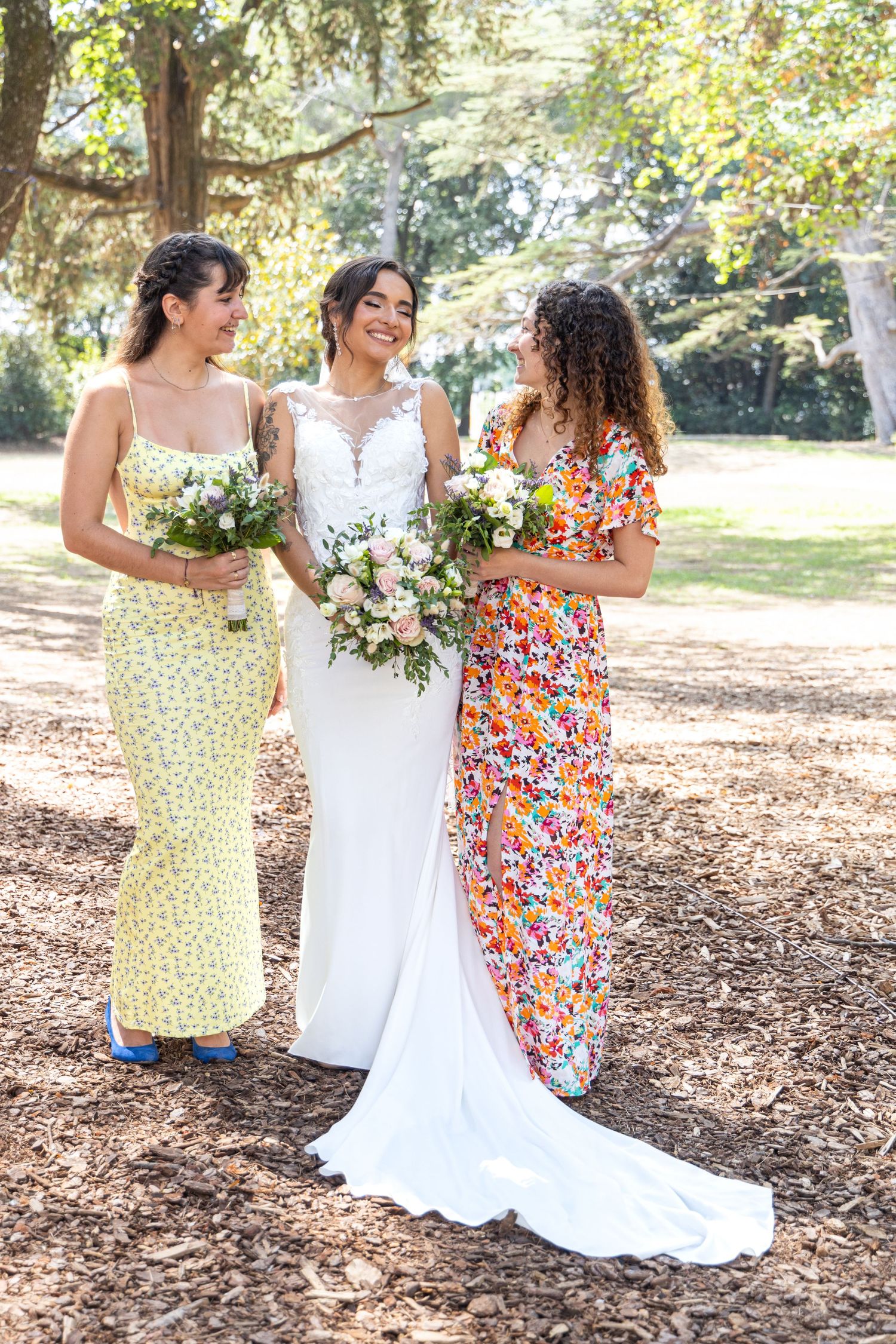 Bridesmaids in colorful floral dresses pose with bride in white gown under trees.