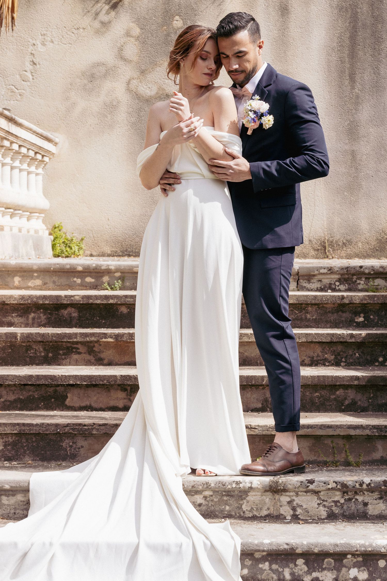 Elegant bride in flowing white gown standing with groom on stone steps.