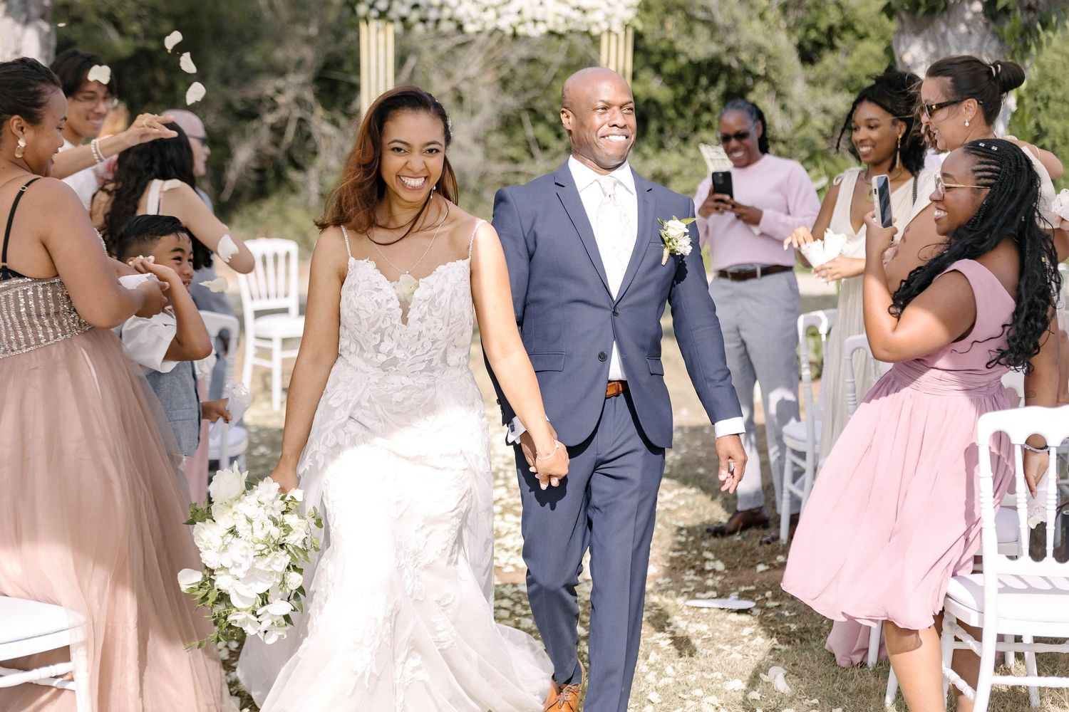 Wedding party celebrating with guests on an outdoor venue decorated with white chairs and flowers.
