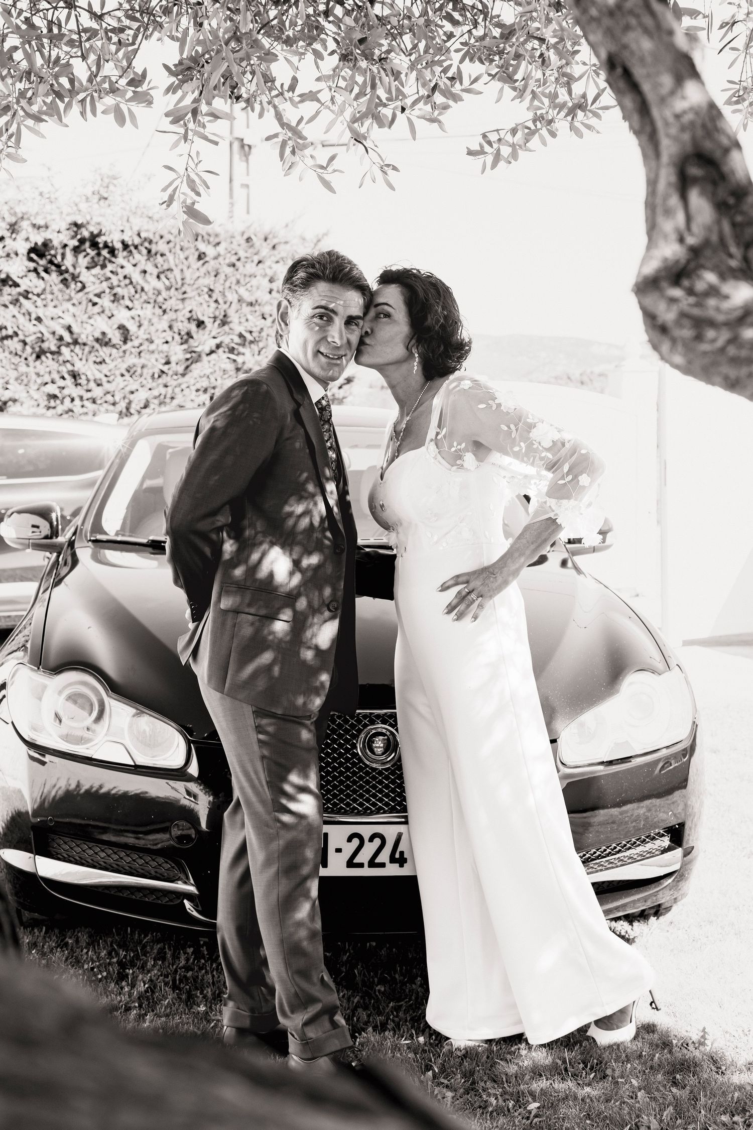 Black and white wedding photo captures playful moments between couple near classic car.