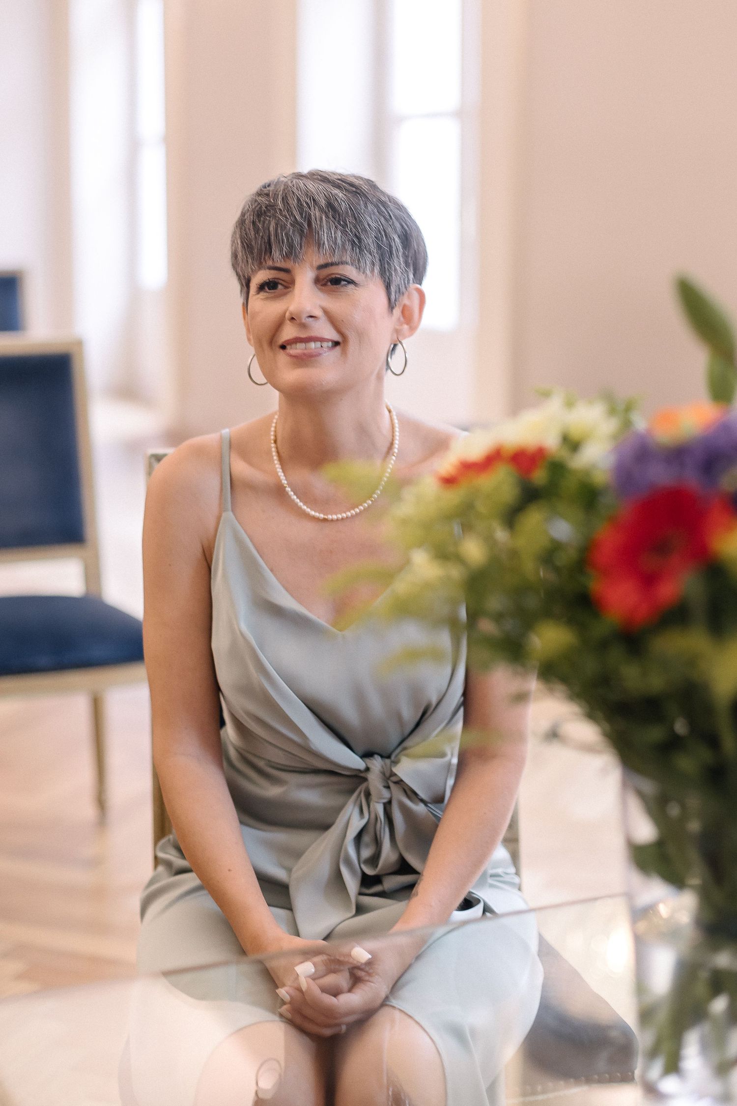 Wedding attendant in silver dress sits elegantly among floral arrangements.