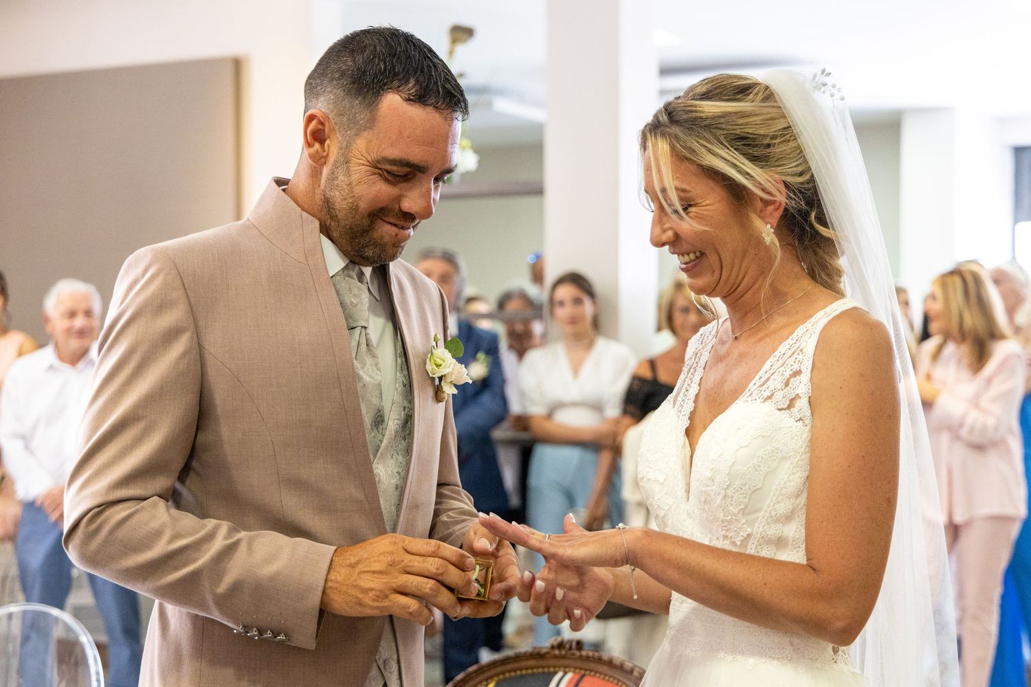 Couple exchanges rings during indoor wedding ceremony with smiling guests watching.