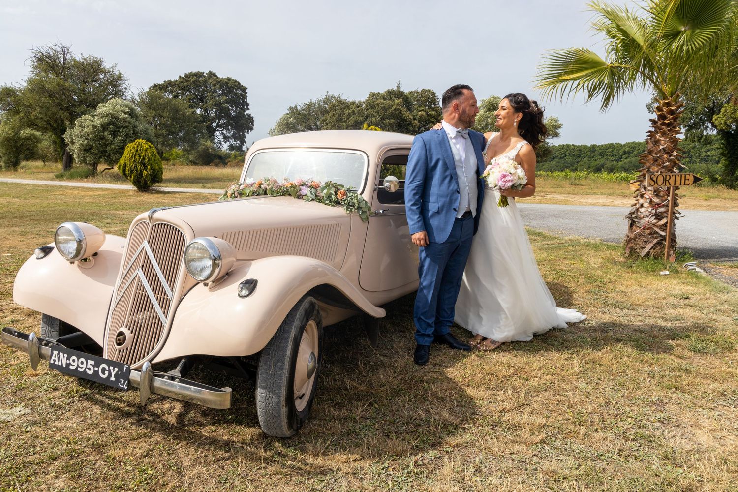 Vintage white car serves as romantic backdrop for wedding couple's portraits in rustic setting.