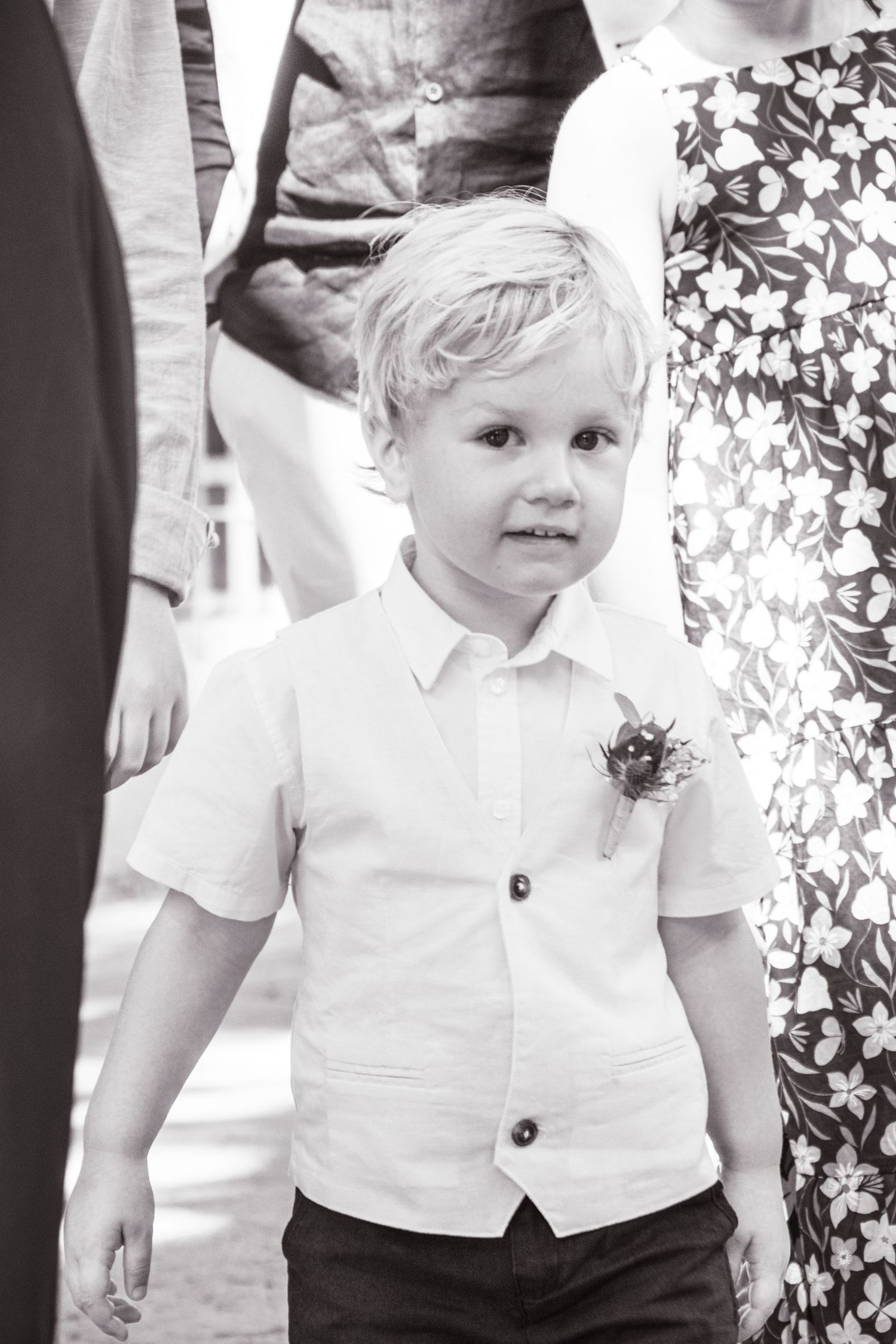 Young child in white dress shirt and vest looks on during wedding celebration.