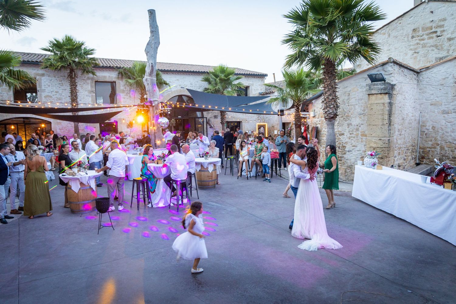 Wedding guests dance on an outdoor patio decorated with string lights and palm trees.
