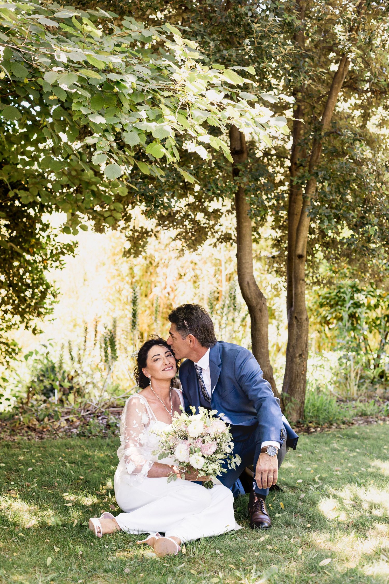 Wedding couple relaxes on grass under trees during their outdoor photo session.