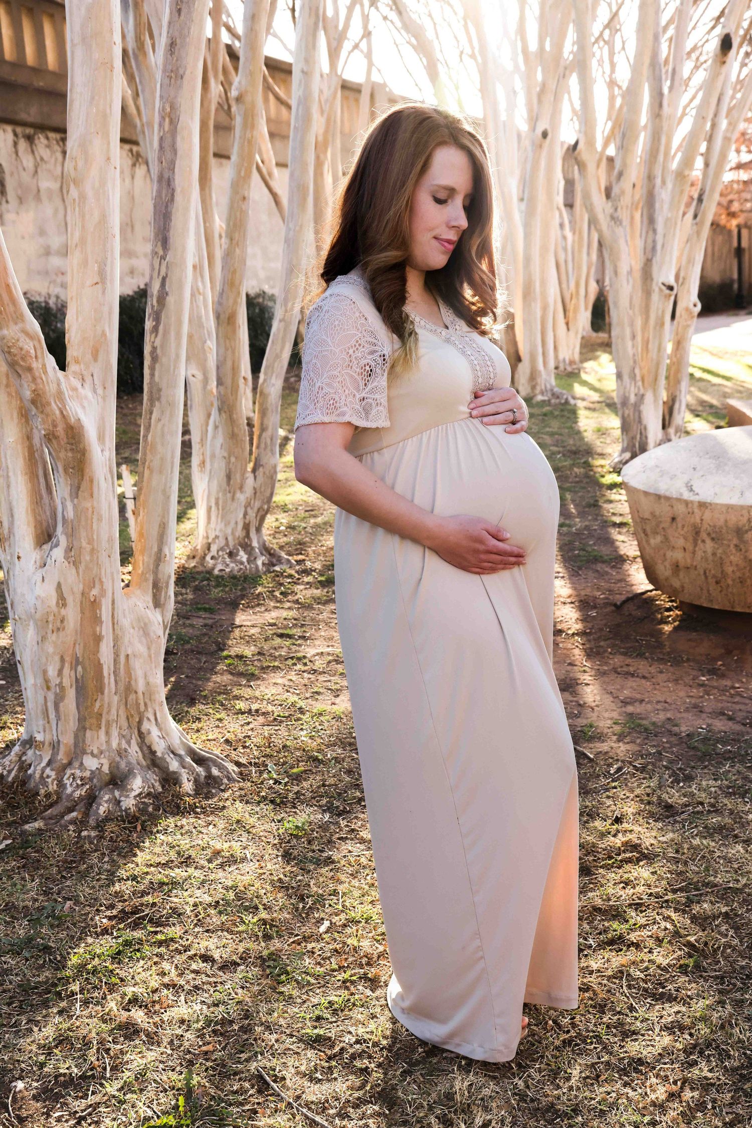An expectant mother in a cream lace dress poses among white-barked trees in Abilene, Texas.