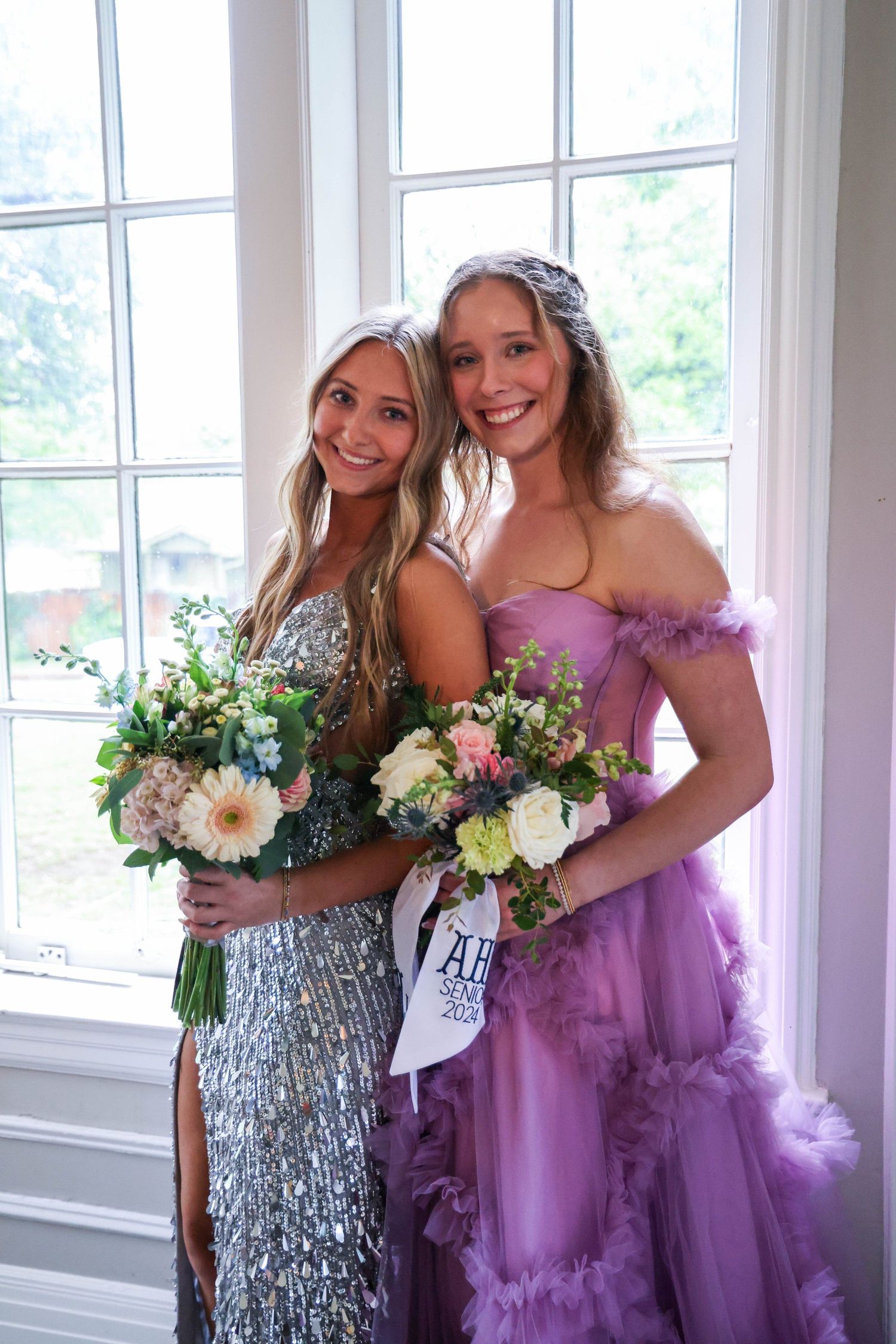 Two bridesmaids in sparkling and purple dresses pose with floral bouquets by a window in Abilene, Texas.