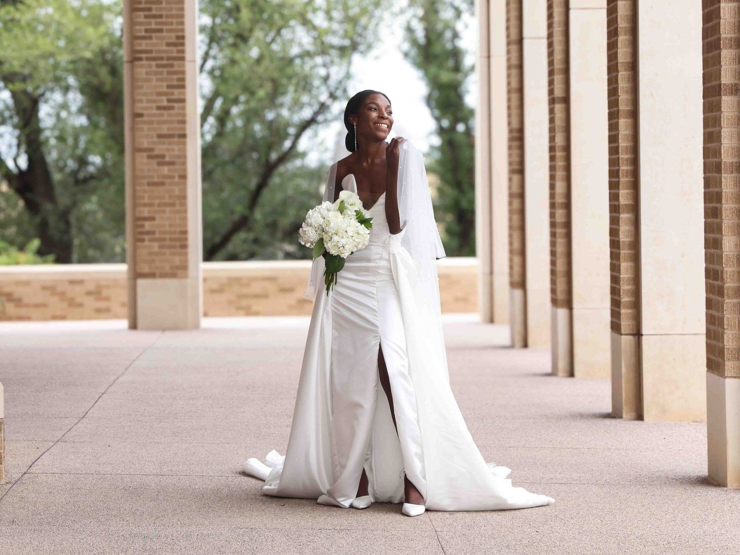 A bride in an elegant white gown poses between brick columns with a bouquet in Abilene, Texas.