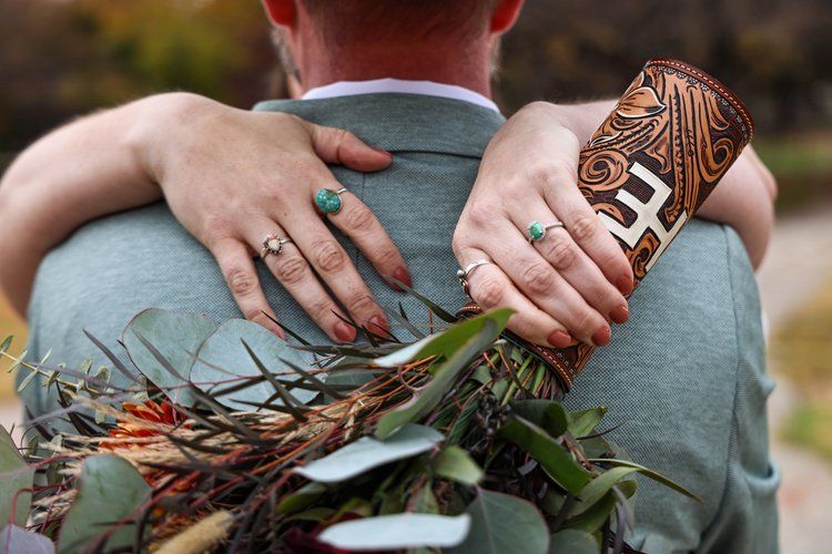 Close-up of hands wearing wedding rings holding a decorated leather bouquet wrap in Abilene, Texas.