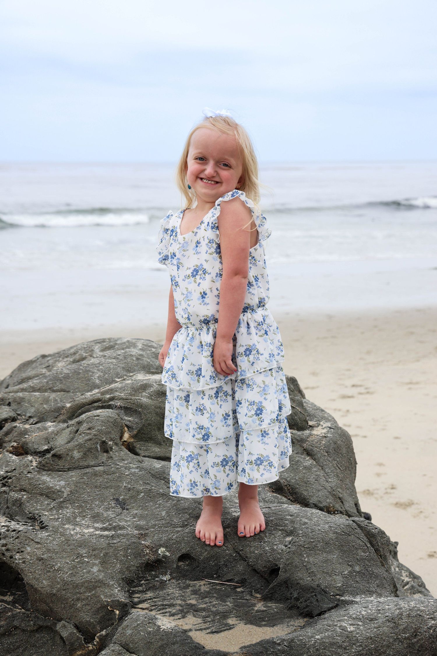 Little girl in floral dress stands on beach rocks with ocean waves behind her.