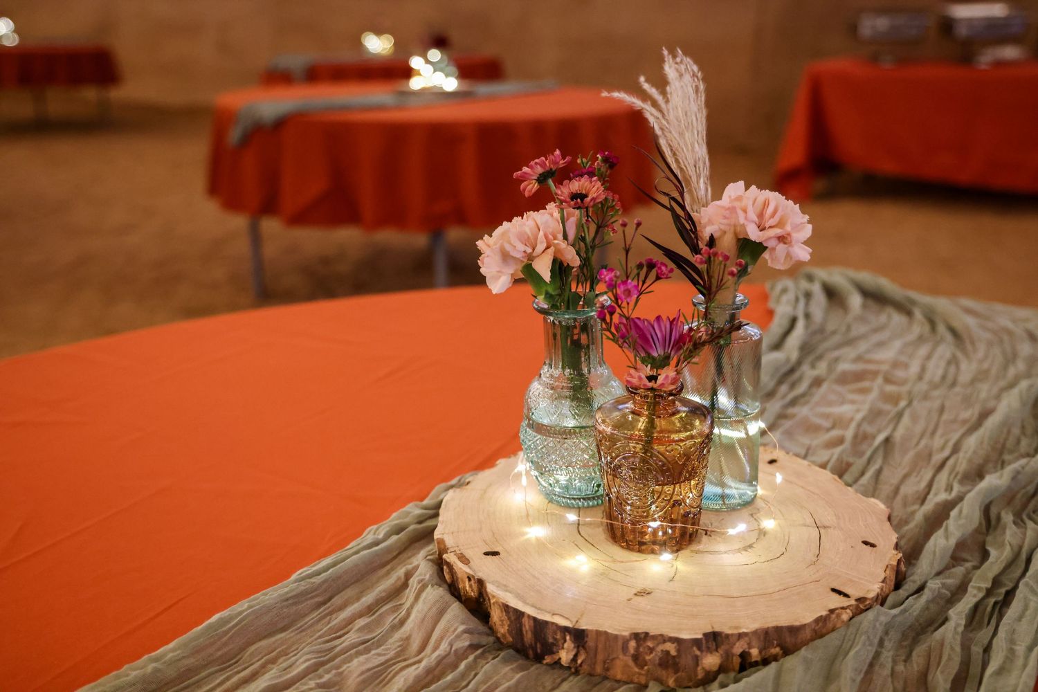 Photograph of rustic wedding centerpiece with pink flowers in mason jars on a wood slice with orange tablecloths in Abilene, Texas.