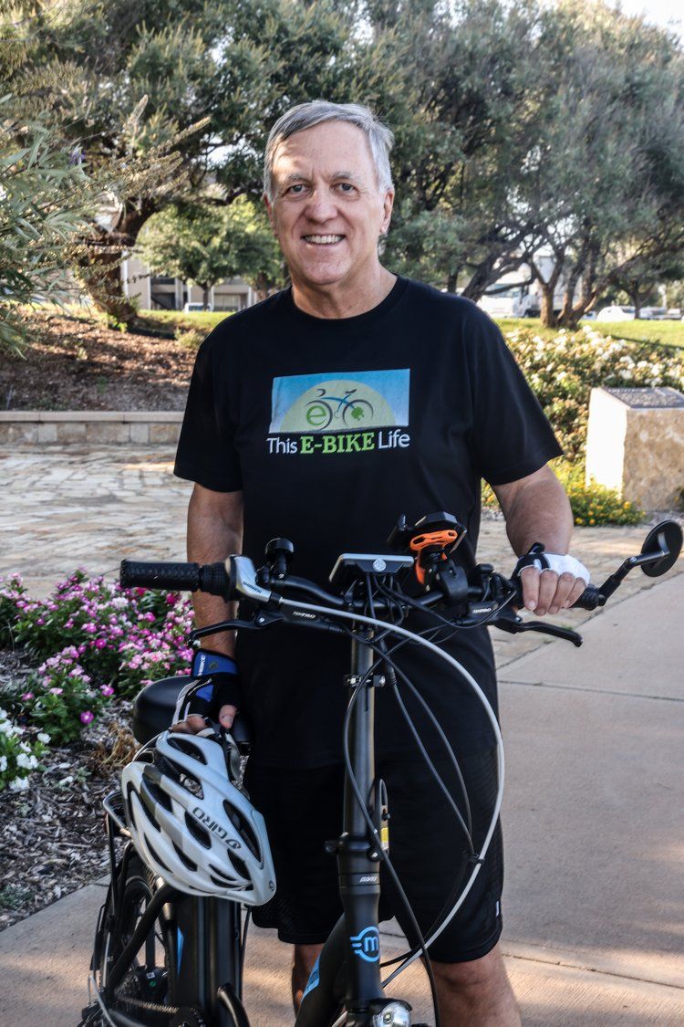 Cyclist in a bike-themed t-shirt poses with bicycle on a sunny path with flowers in Abilene, Texas.