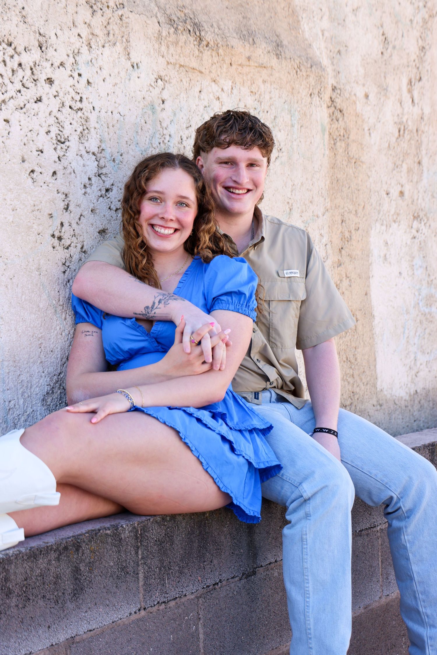 Couple in coordinating outfits sitting close together on concrete ledge against textured wall in Abilene, Texas.
