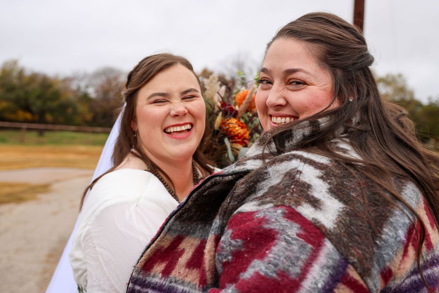 A bride and her bridesmaid laughing together for a photograph outdoors on a cold fall day in Abilene, Texas.