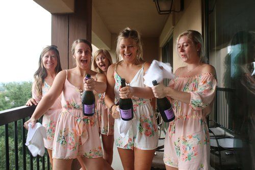 A group of bridesmaids in matching floral robes hold drinks together on a balcony.