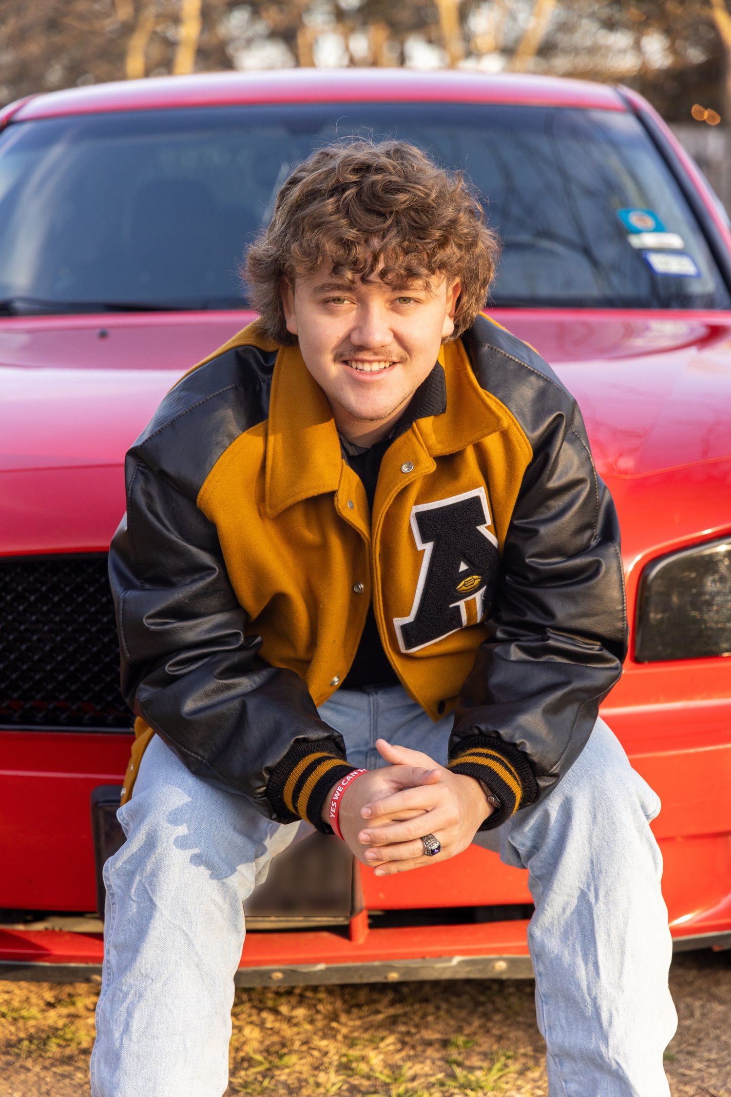 Young person in varsity jacket poses in front of red sports car during Senior session in Abilene, Texas.