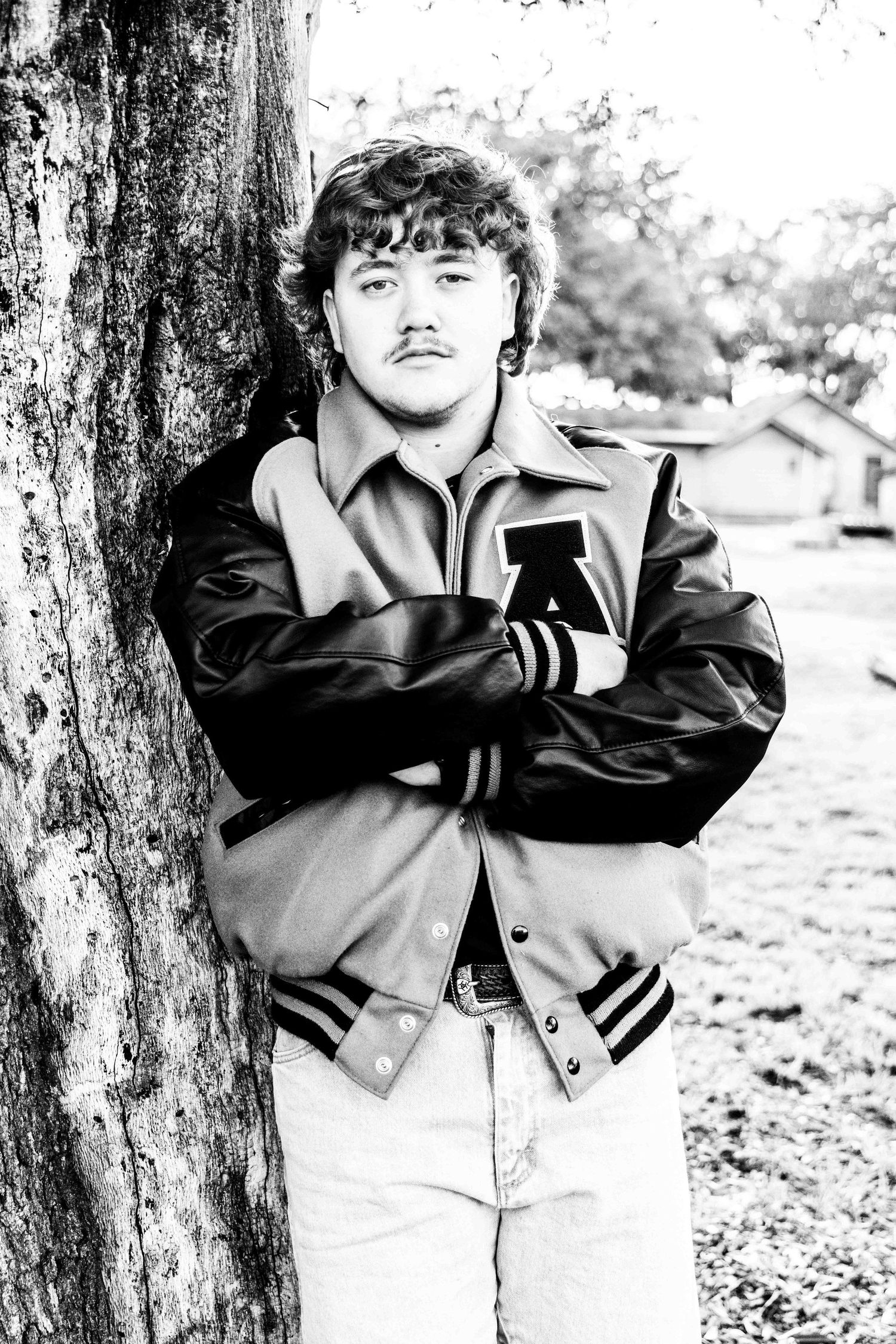 A moody black and white portrait of a young man in a varsity jacket leaning against a tree in Abilene, Texas.