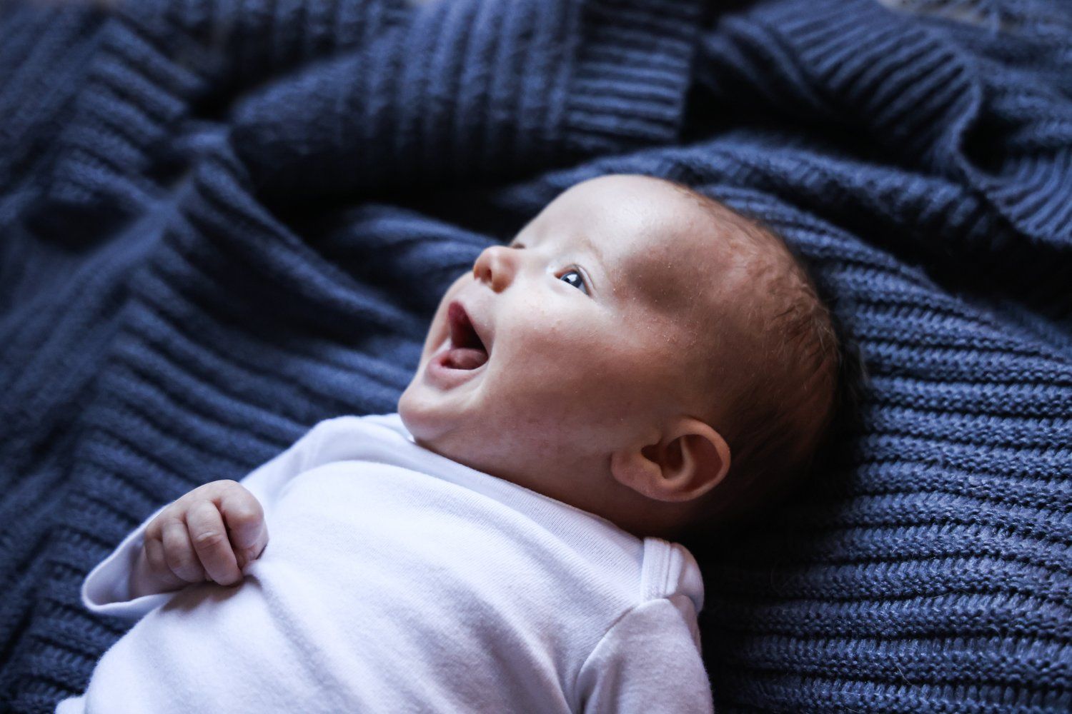 Baby wearing white onesie lies on blue knit blanket looking upward in Abilene, Texas.