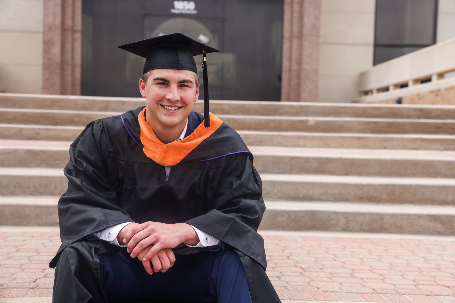 Graduate in cap and gown smiling while sitting on steps in Abilene, Texas.