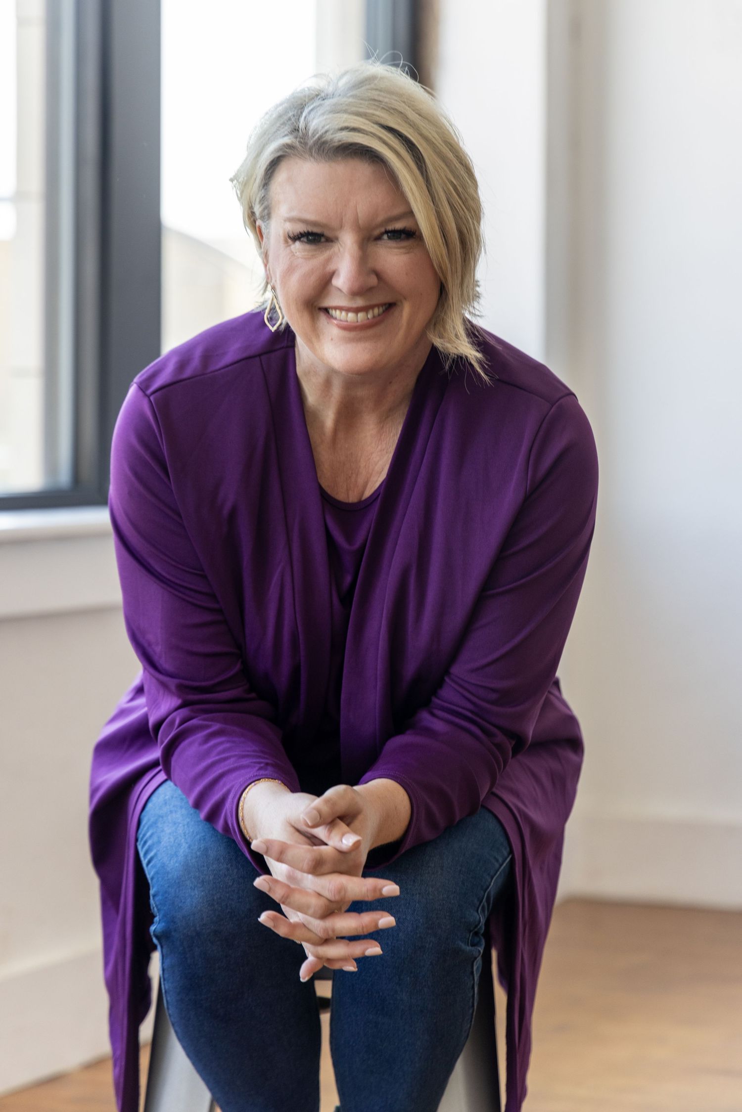 An Abilene woman wearing a purple sweater sits in a bright room with wooden floors and windows for a photography session.