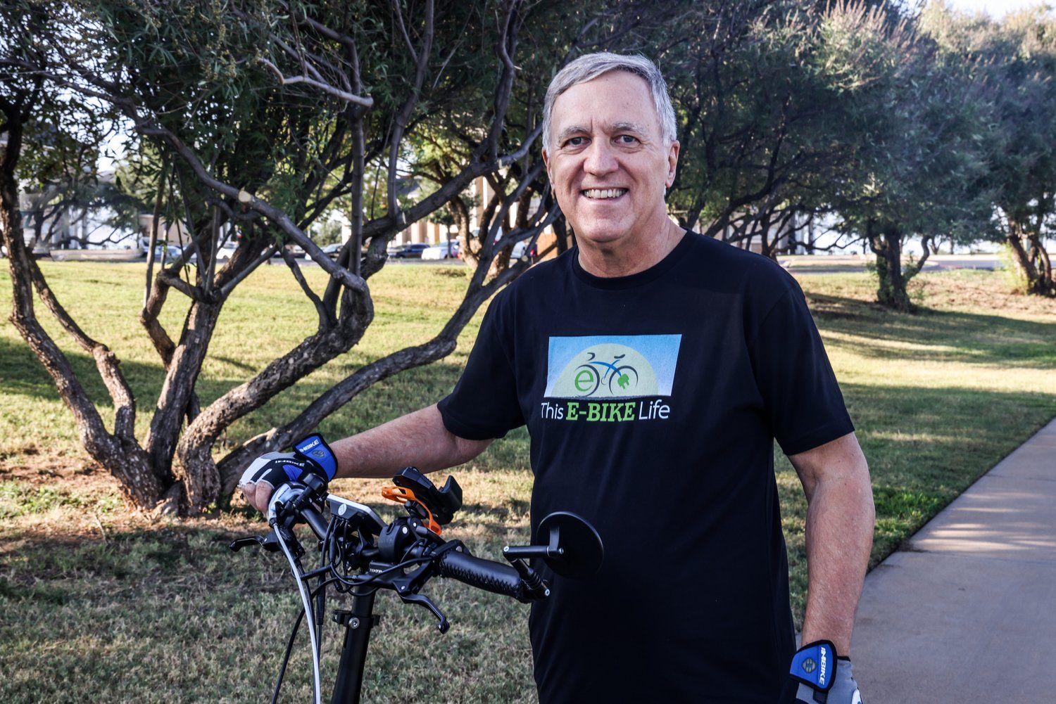 Cyclist in black t-shirt stands with bike on paved trail next to trees in Abilene, Texas.