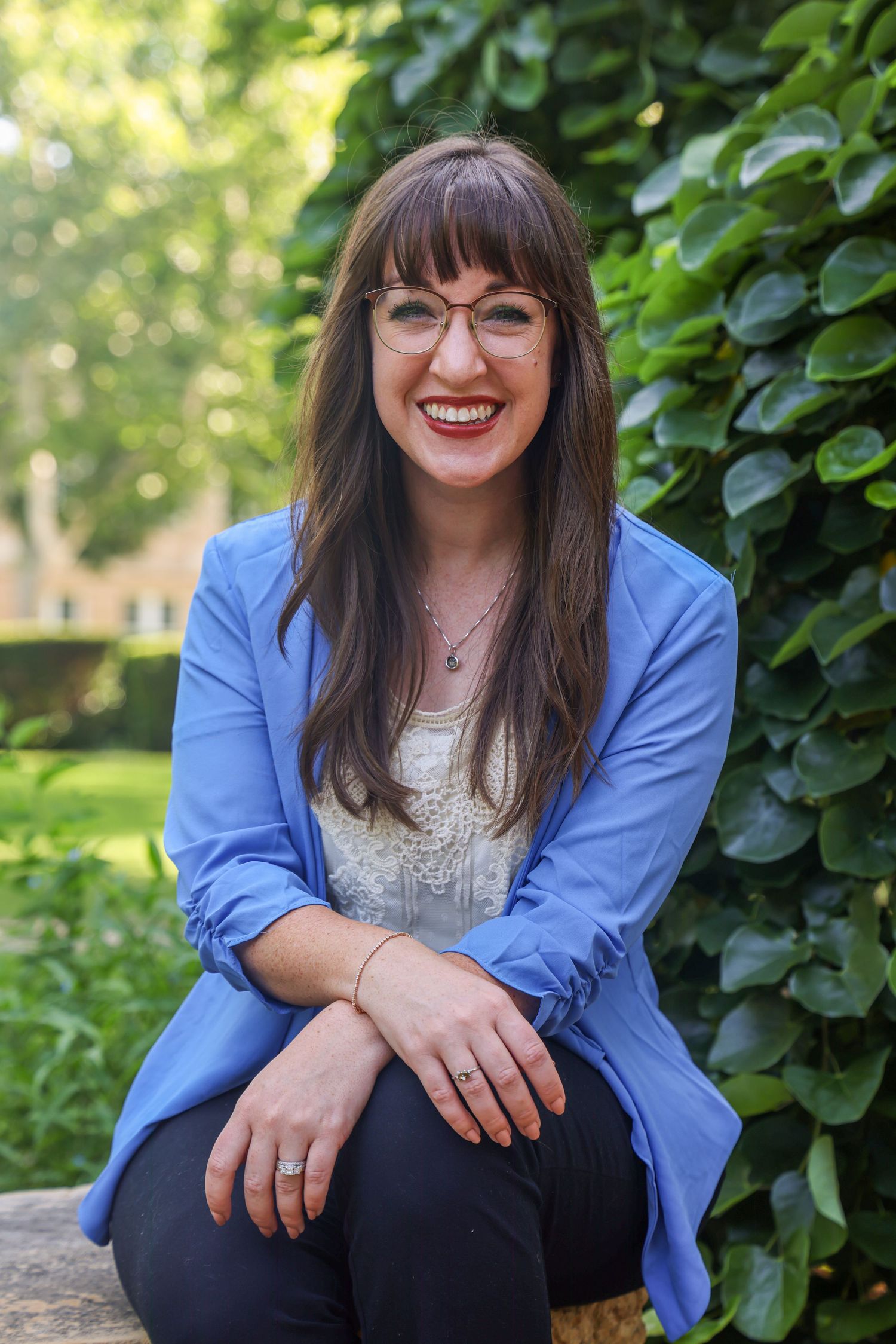 An event speaker in Abilene, Texas in a blue blazer and glasses sits outdoors by greenery, smiling warmly on a sunny day for a Headshot.