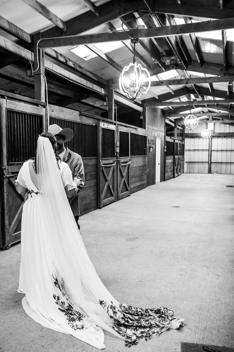 Black and white photo of couple embracing in rustic barn with elegant lighting fixtures.