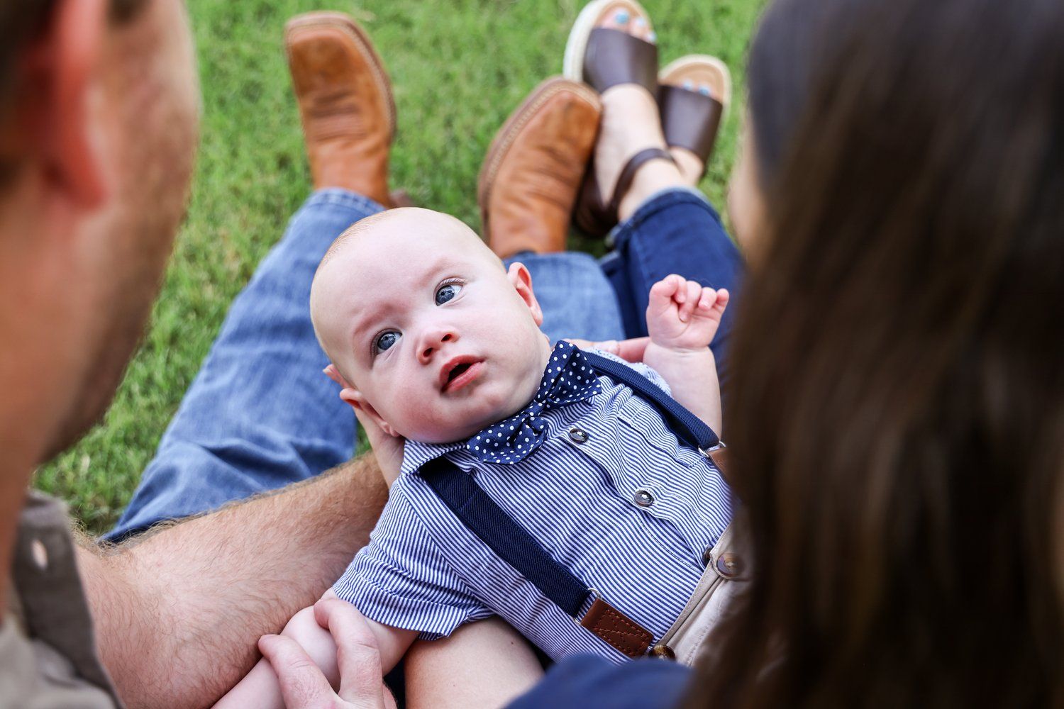 Baby in suspenders and denim lying on grass between two adults in Abilene, Texas.