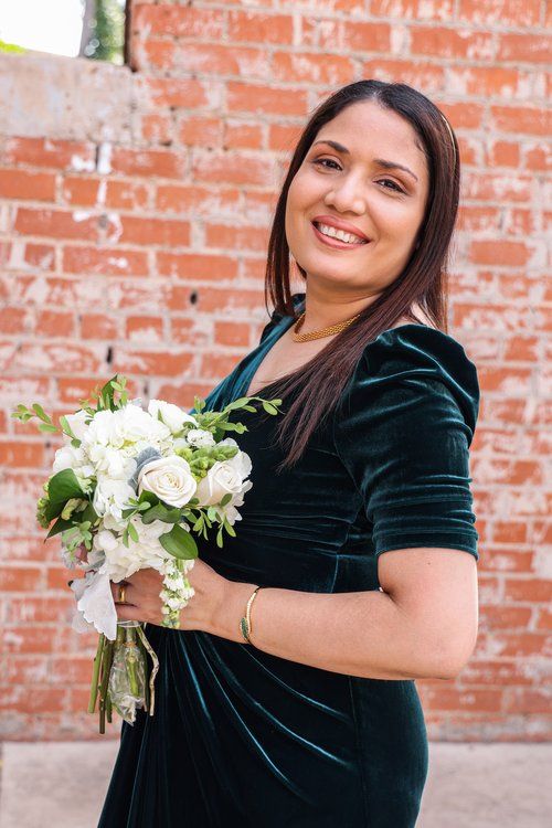 Person in green velvet dress holding white floral bouquet against brick wall.